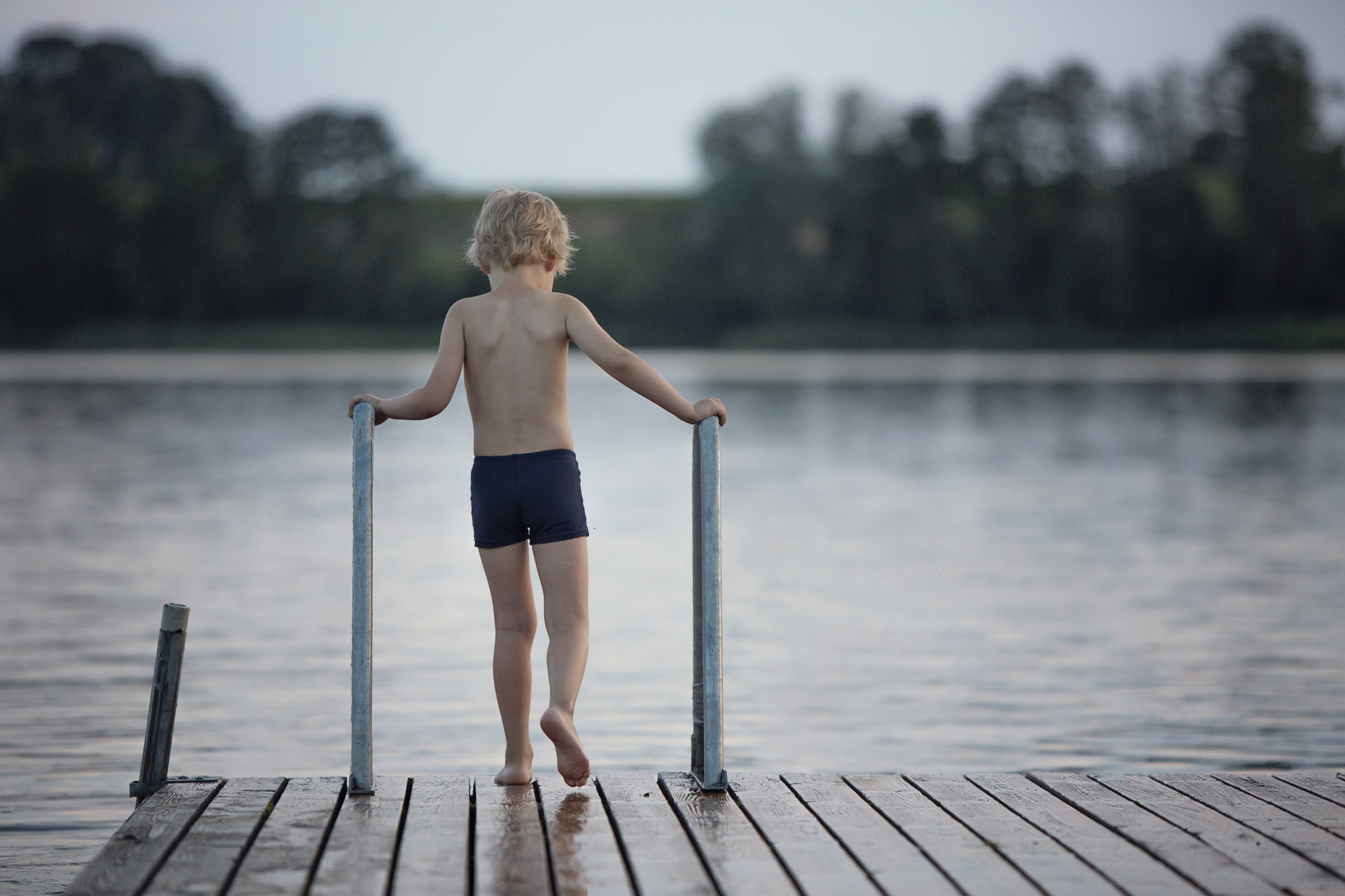 Boy in dark blue swim trunks standing on jetty on a lake.