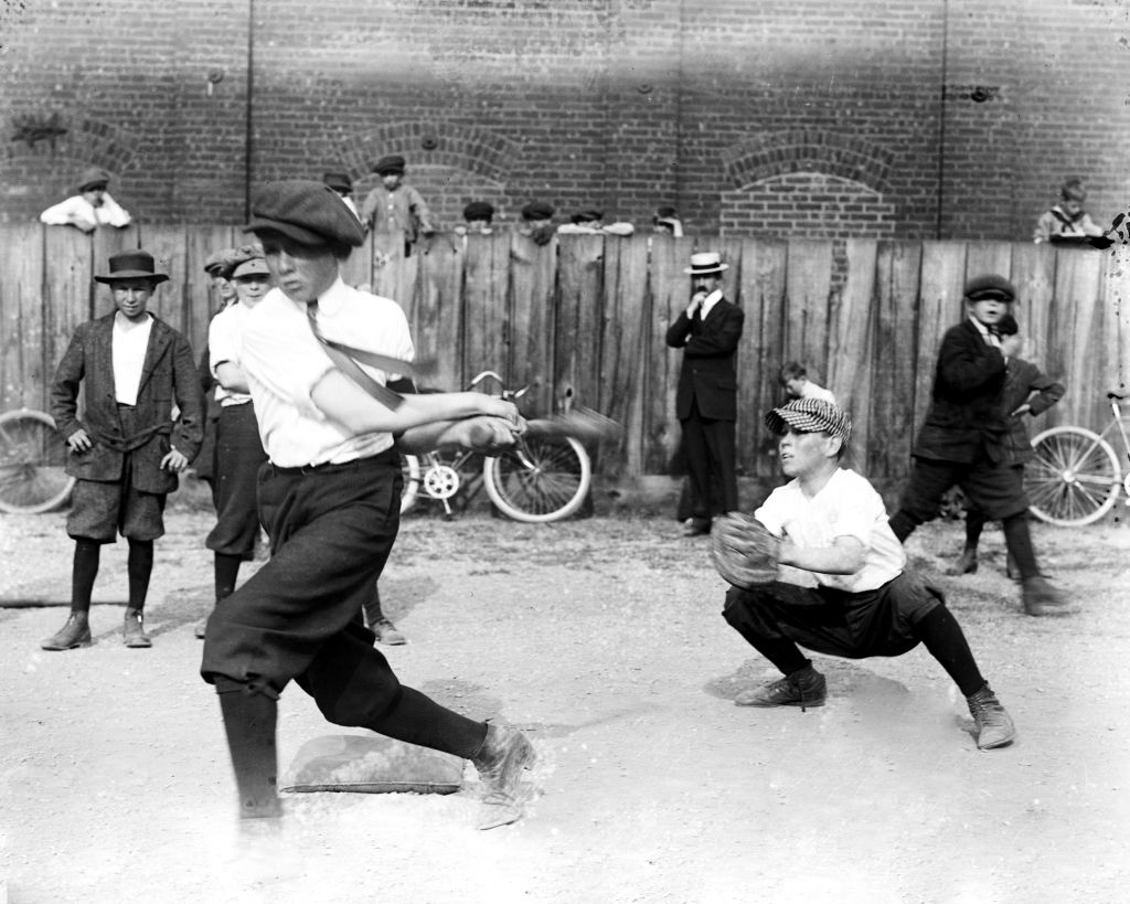 Black and white image of boys playing baseball on a playground ca. 1914. In the foreground, we can see a young boy batting and a catcher behind him. Several other boys watch on in the background.