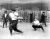 Black and white image of boys playing baseball on a playground ca. 1914. In the foreground, we can see a young boy batting and a catcher behind him. Several other boys watch on in the background.