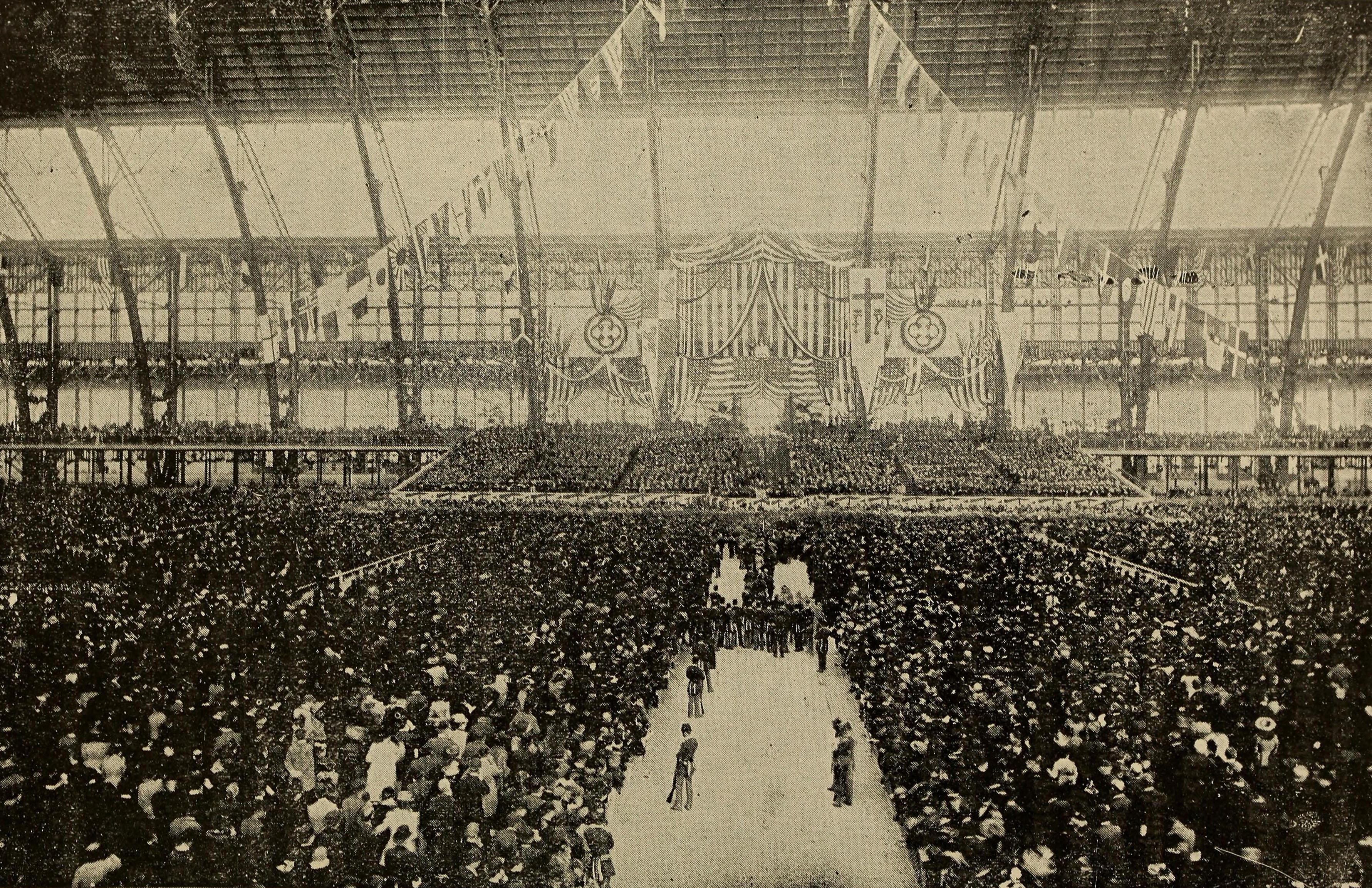 A sepia-toned historic photograph of a huge event. American flags are draped around a main stage in the front.