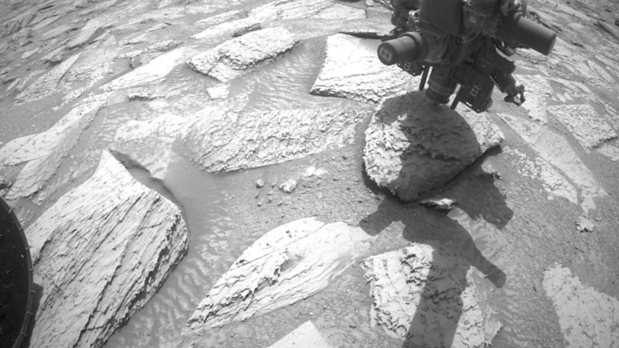 Black and white photo of Curiosity rover's robotic arm drill lifting a stone off the ground on Mars.