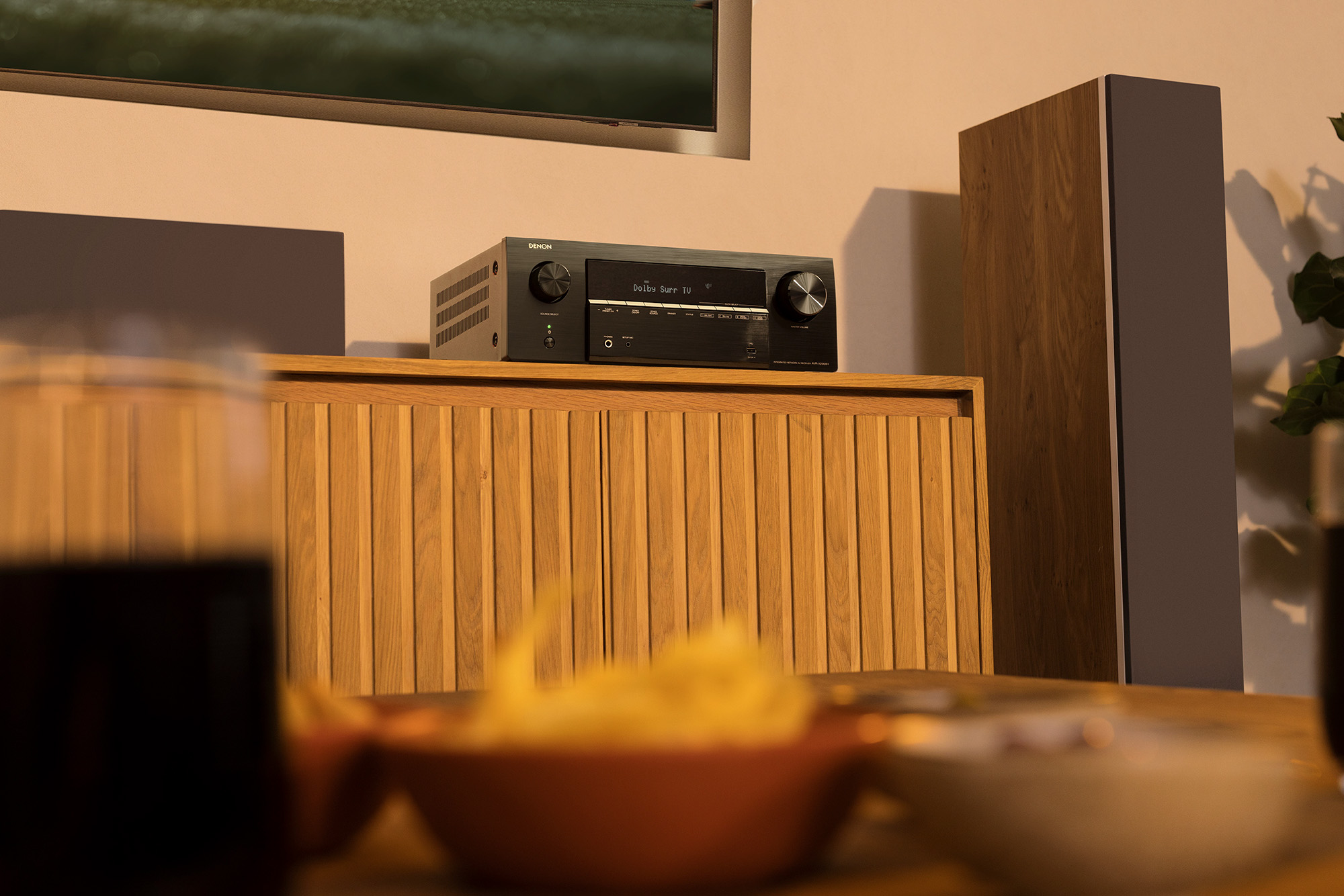 A Denon AVR-X2900 AVR sitting on a wooden midcentury modern A/V cabinet next to a speaker tower.