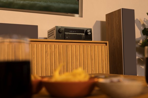 A Denon AVR-X2900 AVR sitting on a wooden midcentury modern A/V cabinet next to a speaker tower.