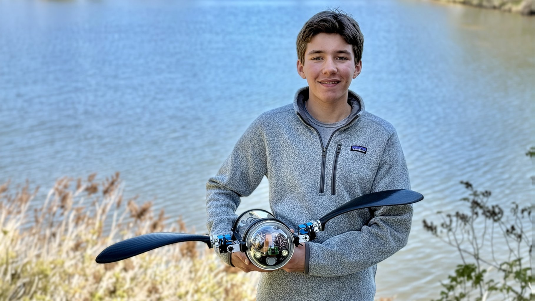 Young white boy in grey pullover holds a robot turtle posing in front of a small pond.