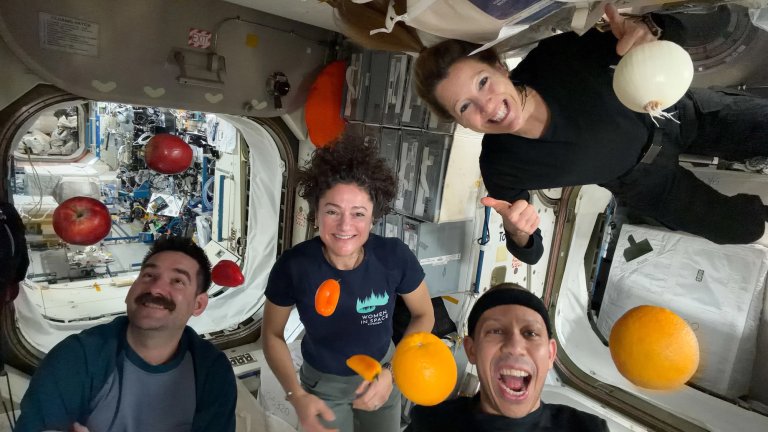 Four ISS astronauts smiling beside floating oranges, peppers, apples, and an onion