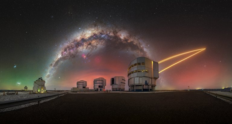 This scene was captured at the Very Large Telescope (VLT) on Cerro Paranal, in Chile’s Atacama Desert, under exceptionally pure observing conditions where the boundary between Earth and sky feels almost seamless. The Milky Way stretches across the sky, with its dense central bulge rising prominently overhead. To the left, the Magellanic Clouds, two neighboring satellite galaxies, are visible to the naked eye from the Southern Hemisphere. Subtle airglow adds another layer to the scene, a natural emission produced by chemical reactions in the upper atmosphere at altitudes of 80 to 100 kilometers. In this image, it reveals a range of colors, with green tones on the left and warmer reddish hues toward the right. In the foreground stands the VLT, operated by the European Southern Observatory. Situated at 2,635 meters above sea level, the site benefits from extremely dry conditions, stable atmospheric layers, and minimal light pollution, resulting in more than 300 clear nights each year. The observatory consists of four primary telescopes, each equipped with 8.2-meter mirrors. One of them, UT4 (Yepun), projects four sodium laser beams into the sky, creating artificial guide stars at an altitude of approximately 90 kilometers. These reference points enable adaptive optics systems to correct for atmospheric distortion in real time, significantly enhancing the clarity and precision of astronomical observations.