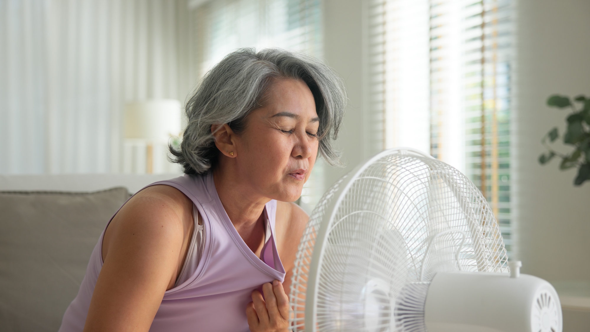 middle age old woman sit at sofa home feel hot flash fever use fan waving