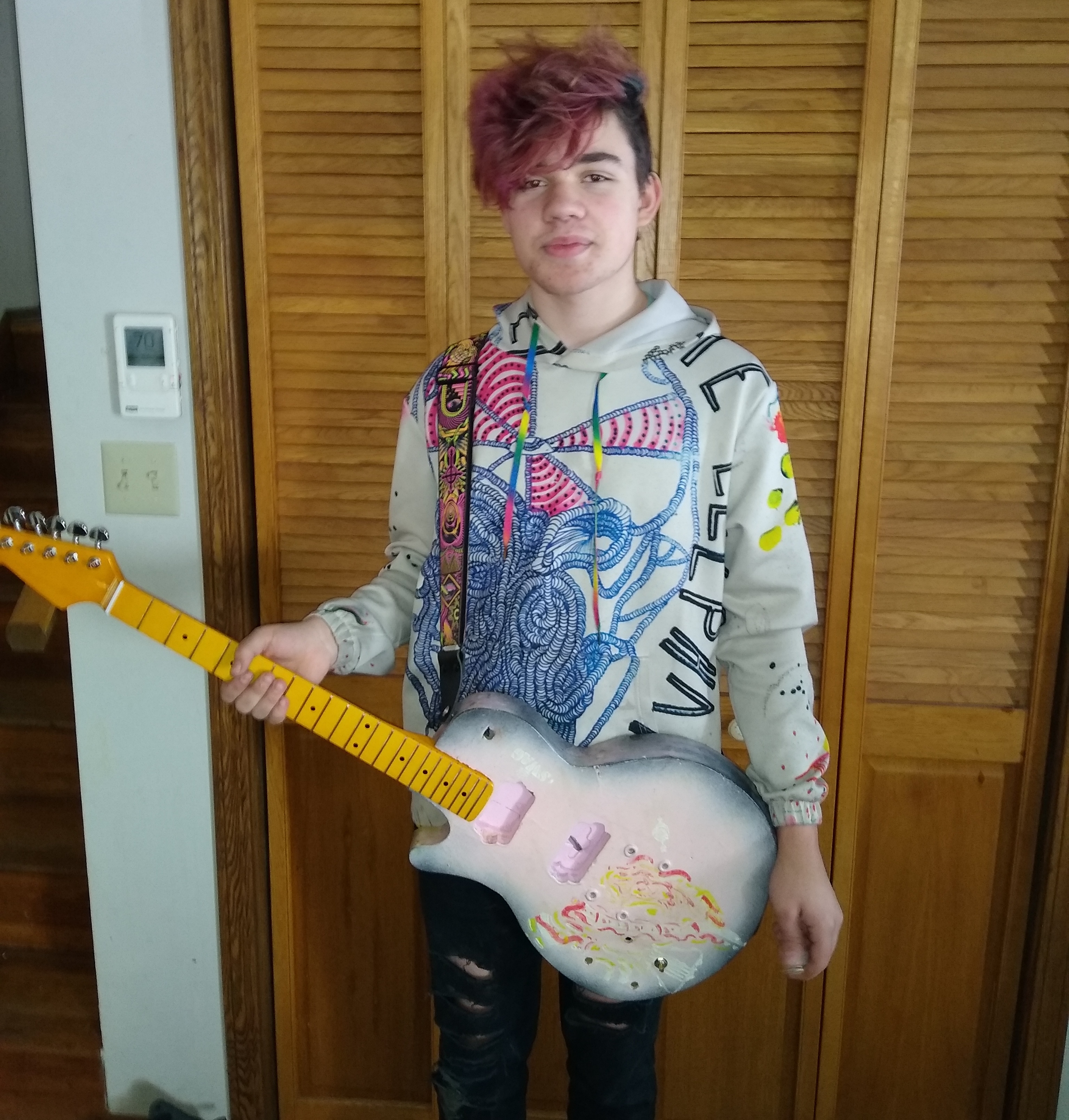 A boy with purple hair wearing a colorful sweatshirt holds a guitar with a blue body and yellow-orange neck.