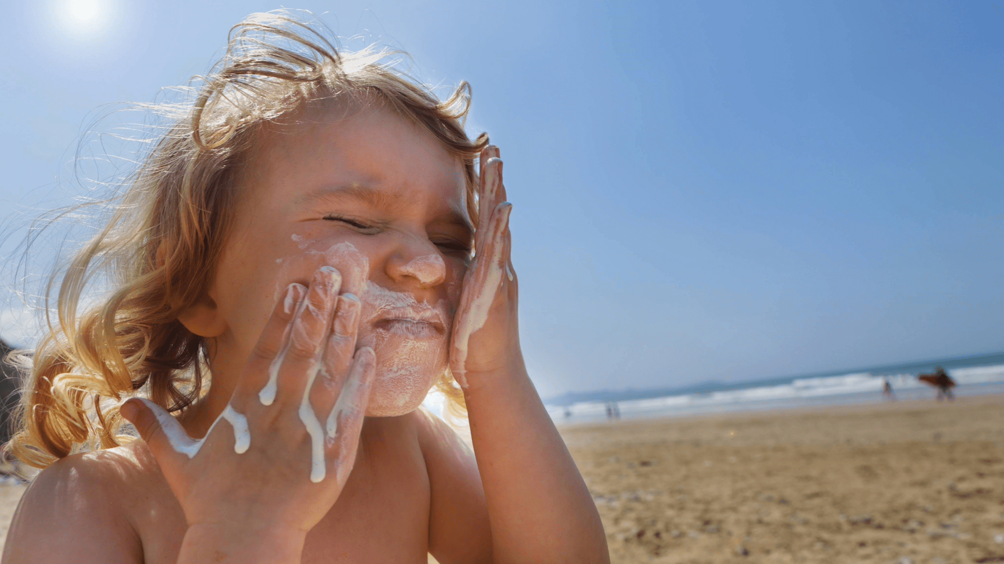 a young child sitting on a beach smears white sunscreen on her face