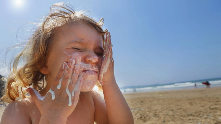 a young child sitting on a beach smears white sunscreen on her face