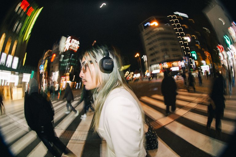 A model with long, flowing bleach-blonde hair, showcasing the Marshall Milton A.N.C. headphones atop his luxurious locks as he walks through the streets at night.