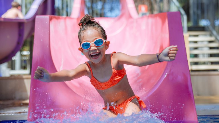 Happy laughing little girl slides down on a pink water slide in outdoor swimming pool on a hot summer day. She's wearing goggles and a orange bikini.