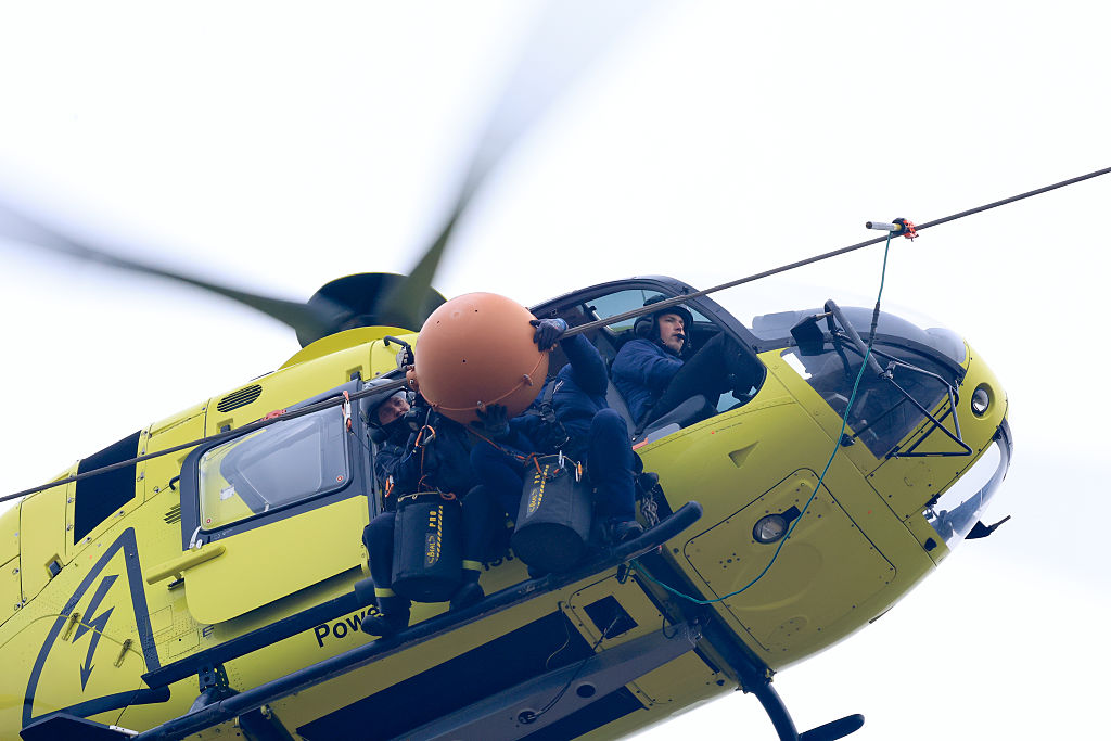 08 March 2026, Achim, Langwedel: Before a power line is dismantled, warning spheres are removed from a power line from the air over highway 27, which is closed for this purpose. A helicopter holds position in the immediate vicinity of the spheres while two fitters sitting on a skid of the aircraft unscrew the orange-colored round bodies. The conspicuous markings are normally used to draw the attention of rescue helicopters approaching emergency scenes on the highway to the lines. Photo: Christian Butt/dpa (Photo by Christian Butt/picture alliance via Getty Images)