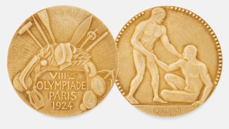 a gold medal from the 1924 olympics in paris. the text on one side reads VIII OLYMPIAD PARIS 1924 and has a rugby ball, discuss, and a harp etched in. the other shows a winner reaching down to help up a fallen competitor over the five interlocking olympic rings