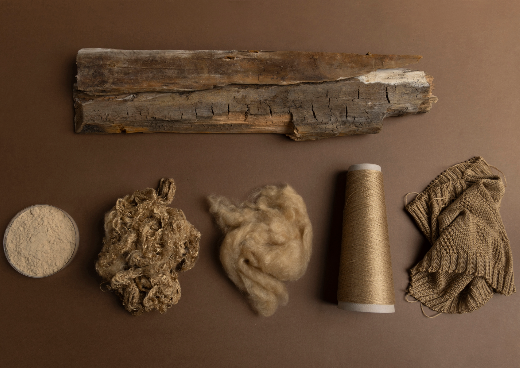 the materials used to make a dress laid out on a brown background. a piece of wood is on top, with wood pulp, fiver, yarn, and he fabric below
