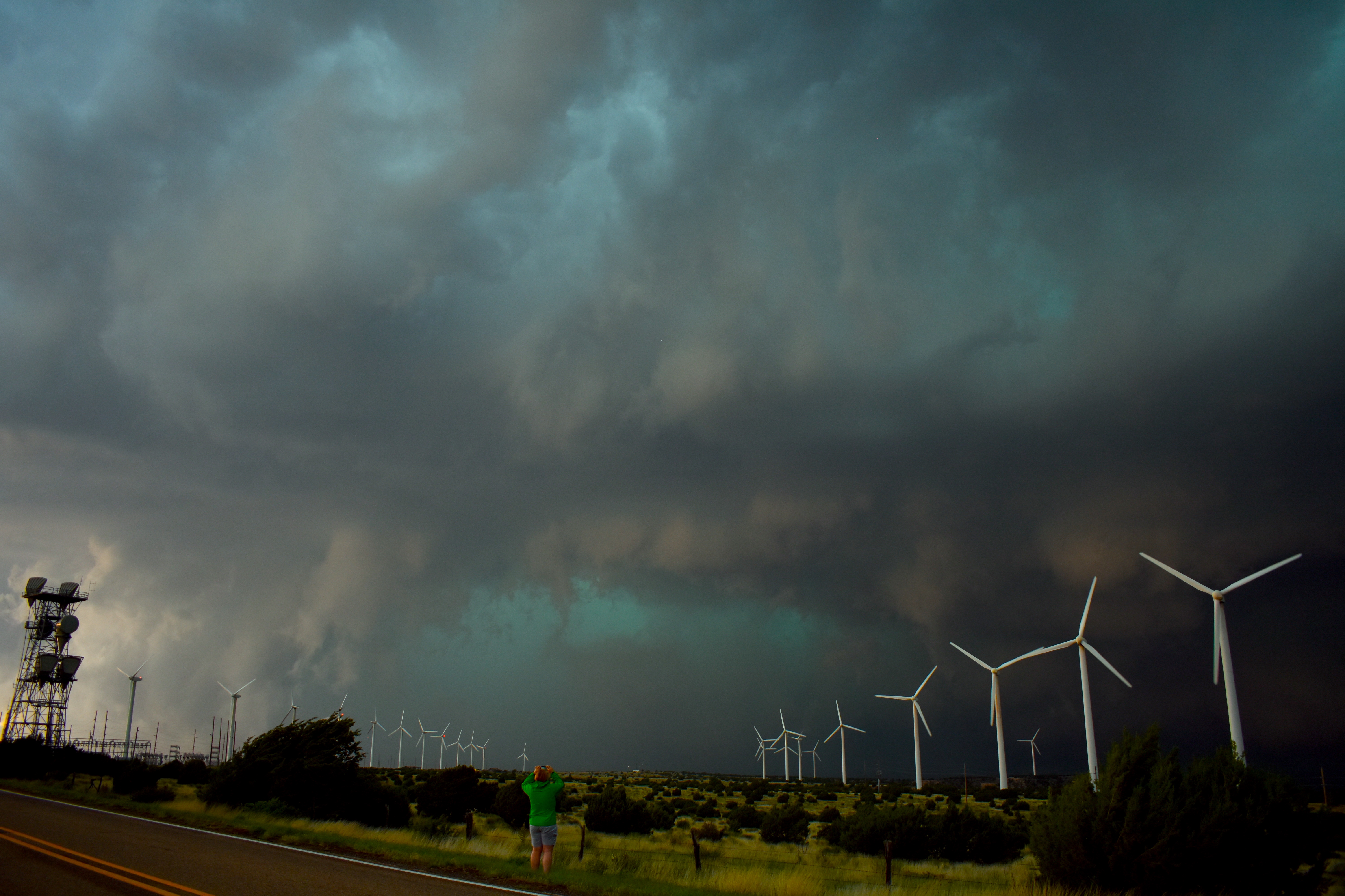 A person looks at a storm forming off the side of the road. There's several wind turbines on the rural countryside.