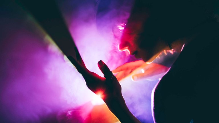 An atmospheric photograph shows a silhouette of a person interacting with a dramatic play of light and shadow. The person’s face and hands are partially illuminated by a bright, warm light source that cuts through a hazy, purple-tinted atmosphere filled with smoke or mist. The composition is abstract and moody, emphasizing the sensory and ethereal nature of light and color.