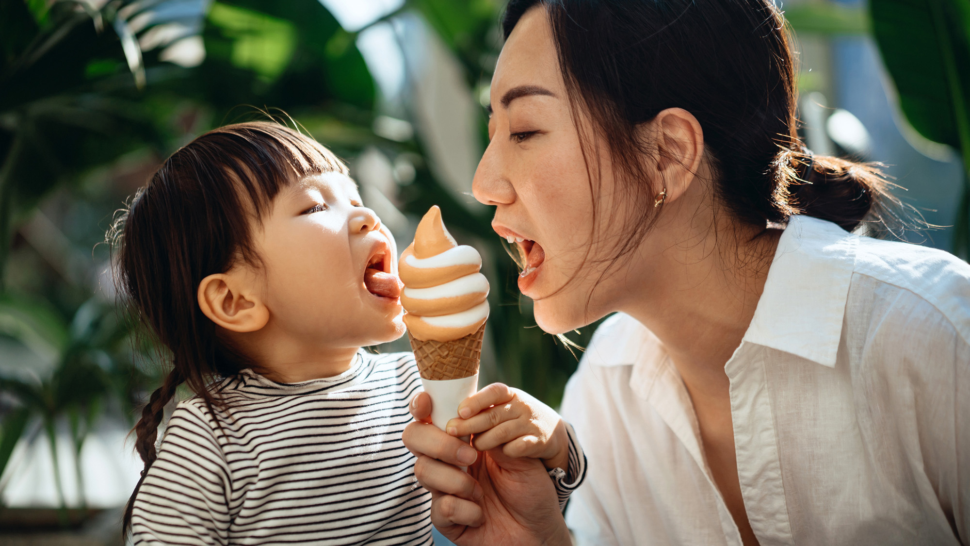 Loving young Asian mother and daughter sharing ice-cream cone joyfully together in outdoor cafe against green plants and sunlight. Sharing mother-daughter moments. Having fun while dining out. Family, food and lifestyle