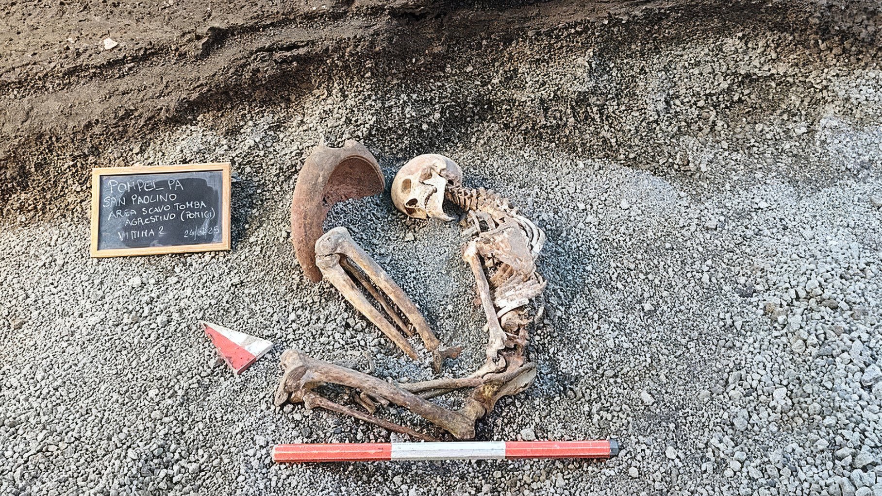 Skeleton in the fetal position on gravel with a mortar near his head. A chalk board has some information on the side.