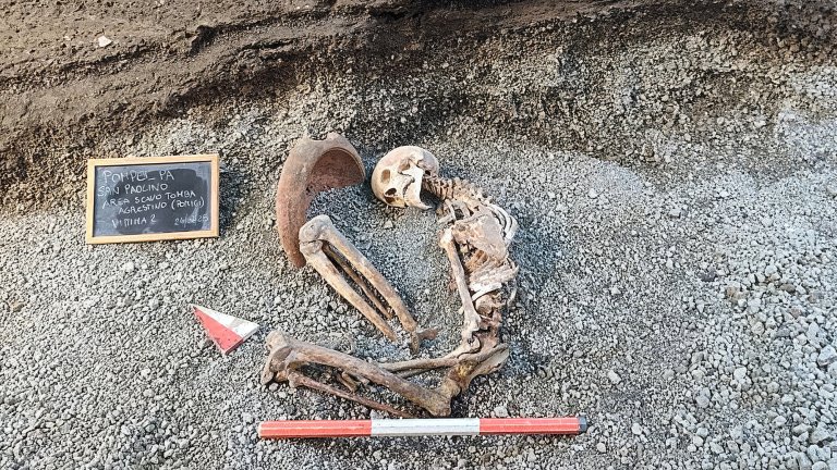 Skeleton in the fetal position on gravel with a mortar near his head. A chalk board has some information on the side.