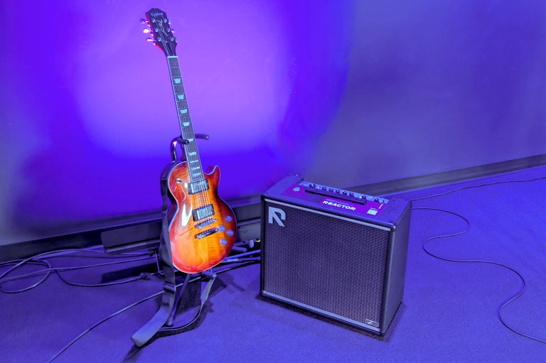 A Positive Grid REACTOR AI practice amp on stage next to a sunburst Gibson Les Paul guitar bathed in purple light