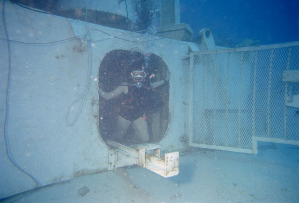 A woman in a scuba gear emerges from an underwater habitat.