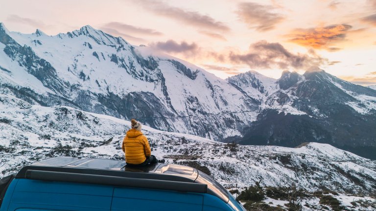 Person in orange puffer jacket sits on the top of a van and looks out at the French Pyrenees mountains.