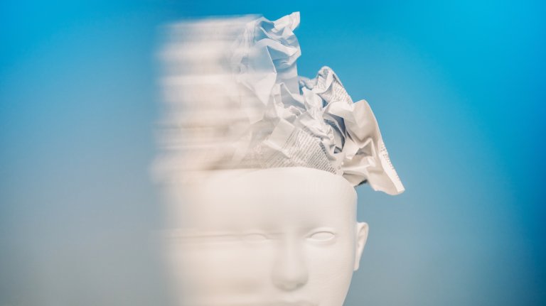 A white stylized head with crumpled paper overflowing from the top is captured with a motion blur effect against a blue background. This conceptual art represents a scattered mind, information overload, and the chaotic nature of overthinking.