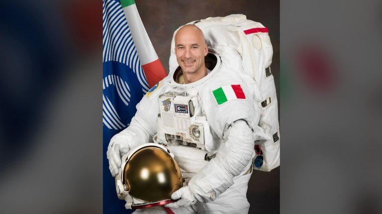 a male astronaut wearing a white space suit with an italian flag on the arm holds his helmet