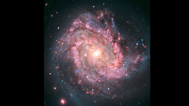 In the composite image, Messier 83, or M83, is shown to have a spiral structure, viewed straight on. At the center is a brilliant white and yellow pool of light. From that light, spiral arms of hot pink cloud corkscrew out in wide, sweeping arches. The galaxy is covered in a faint grey haze, and flecked with red, green, blue, white, and yellow dots.