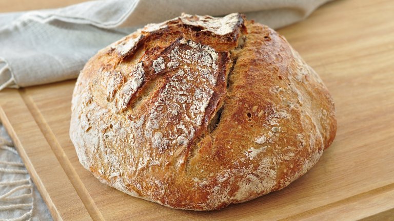Round sourdough bread loaf, with a crispy crust, dusted with flour, resting on a rustic linen cloth on a wooden surface. Home bakery, traditional baking concept.
