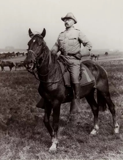 teddy roosevelt on a horse. he is wearing a wide-brim hat and glasses