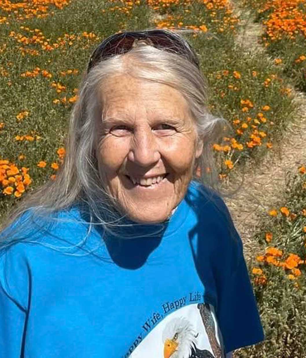 a woman with long grey hair and a blue t-shirt smiles while standing in a field of orange poppies