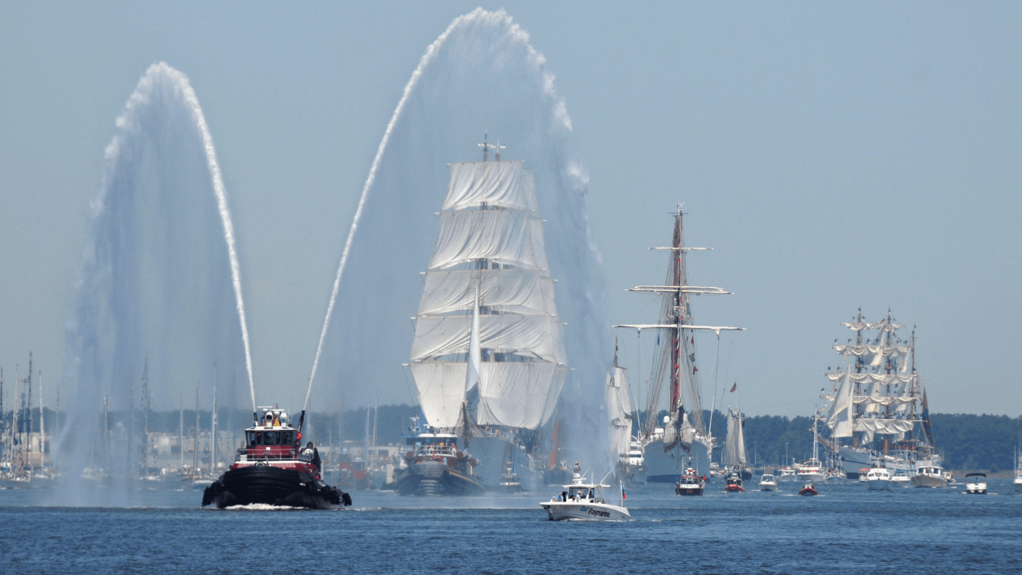 a water canon sprays water while several tall ships sail into new york harbor