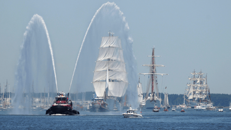 a water canon sprays water while several tall ships sail into new york harbor