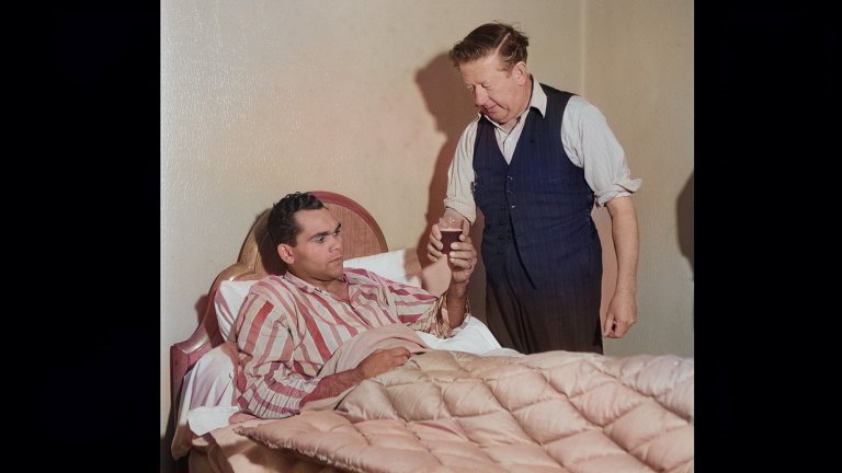 A man in pjs lying down in a small bed takes a glass cup from an older man.