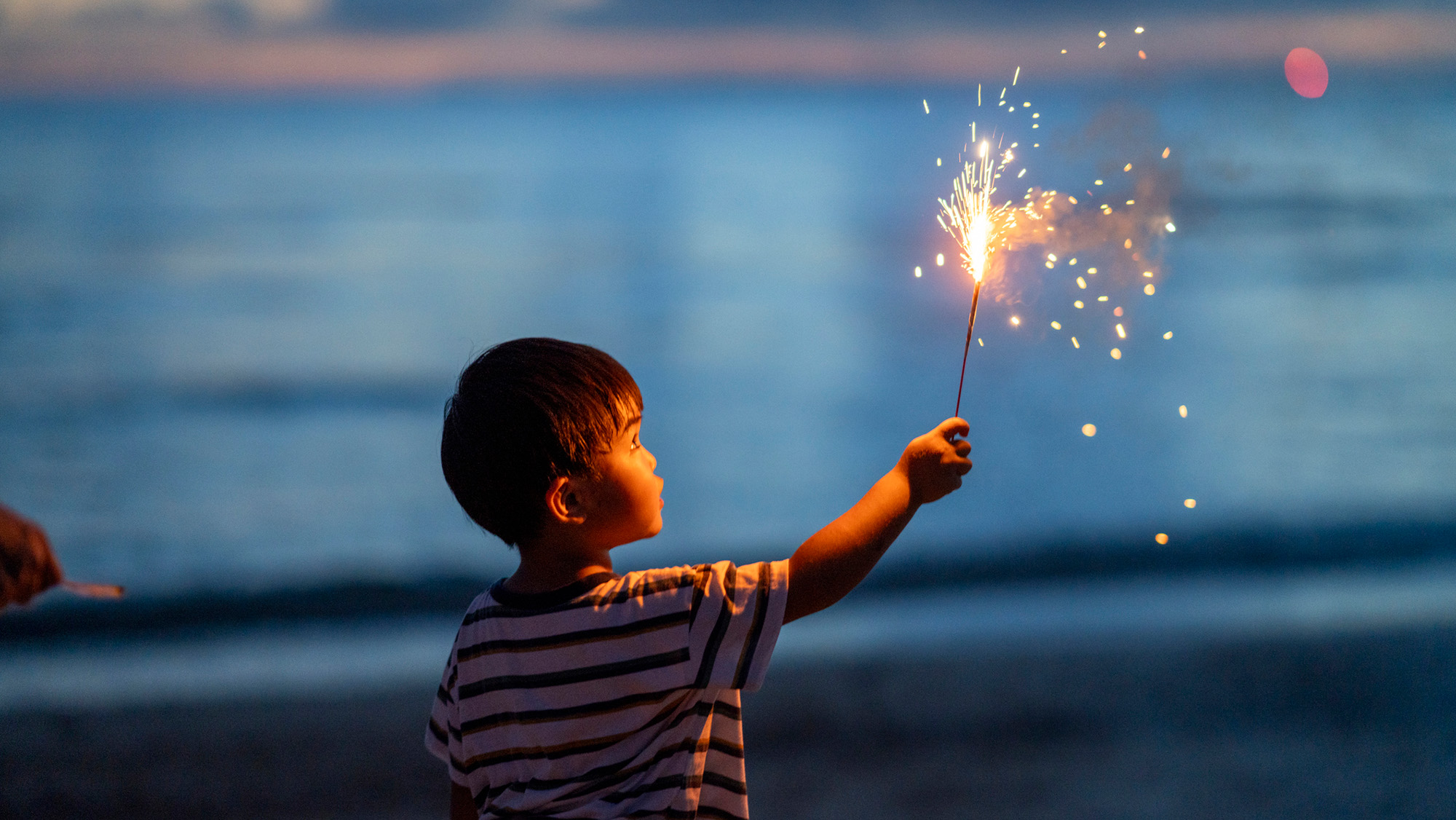 A little boy in a striped shirt holding a sparkler in front of a lakeshore.