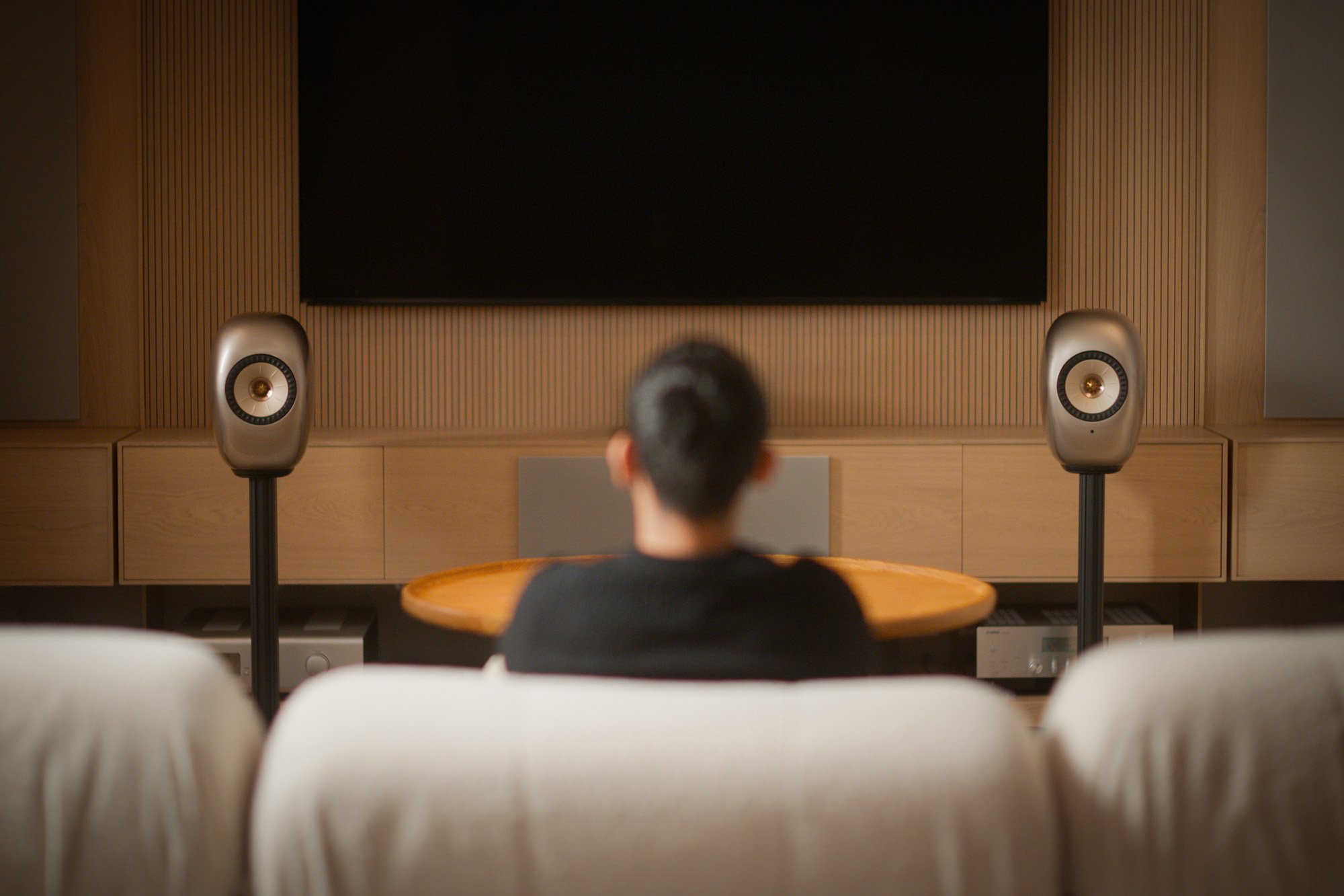 A man sitting on a couch in front of a pair of metallic KEF LS LUXE active all-in-one wireless hi-fi speakers on stands.