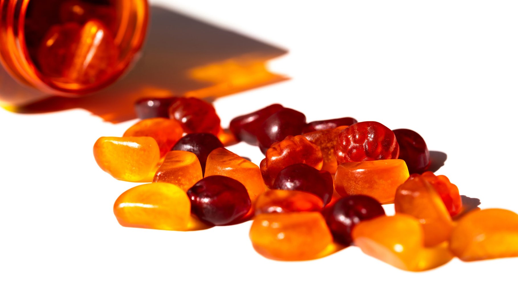 Close-up of assorted red and orange gummy vitamins spilled from a transparent bottle on white background.