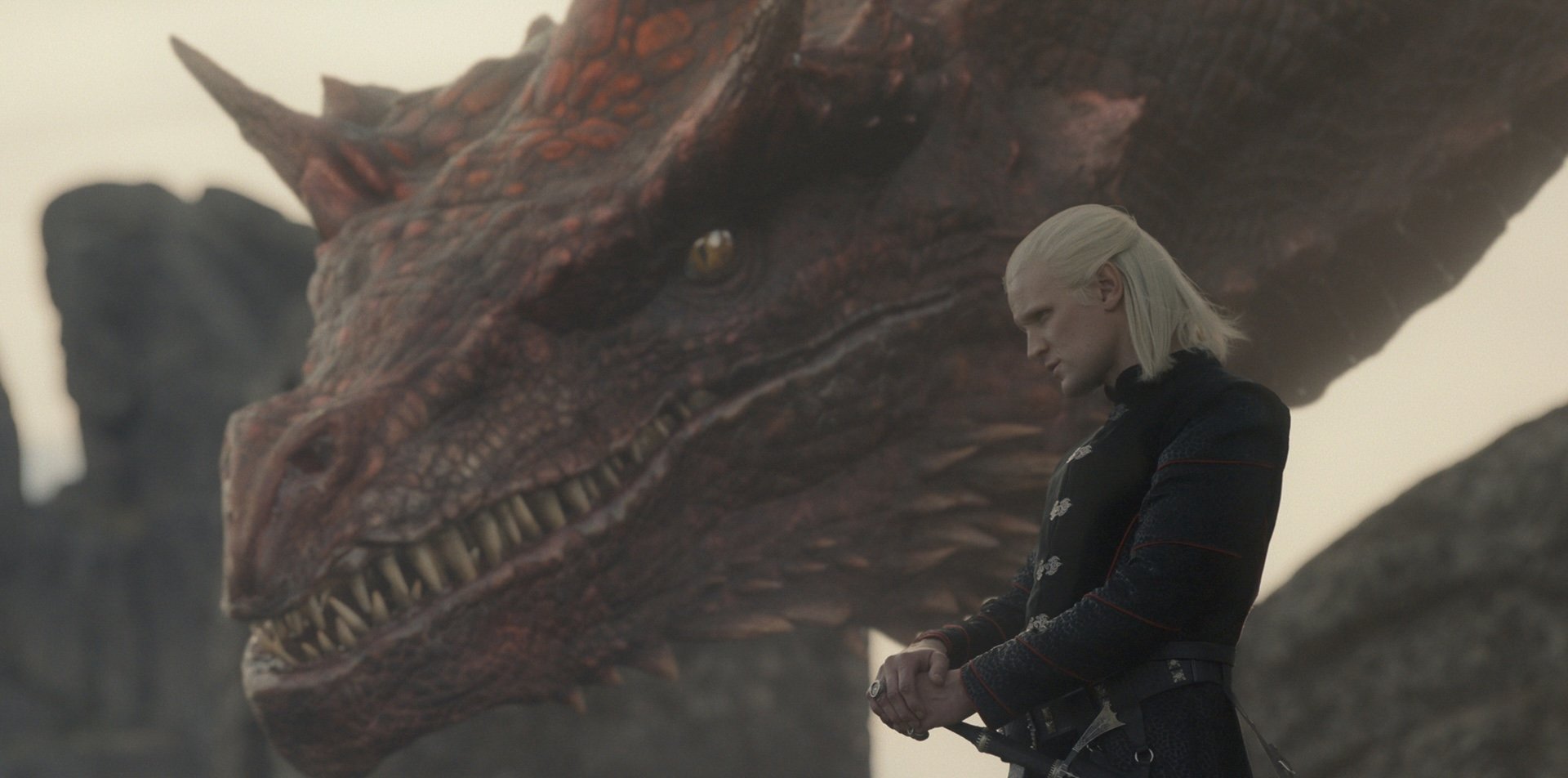 Man with chin length white-blonde hair stands in front of a dragon with a red head.