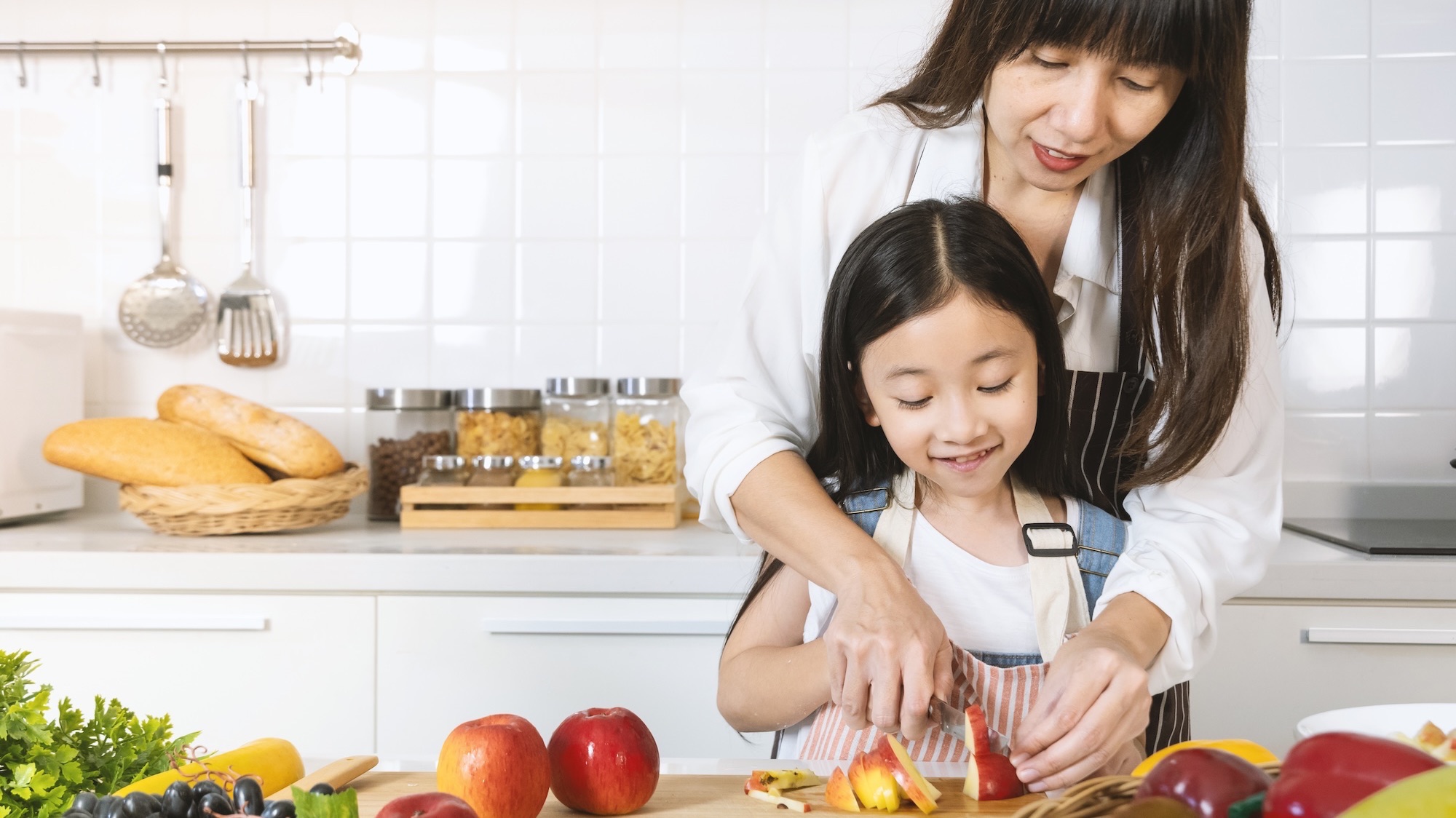 Mother and little daughter using knife to preparing a healthy fruit snack in kitchen at home.