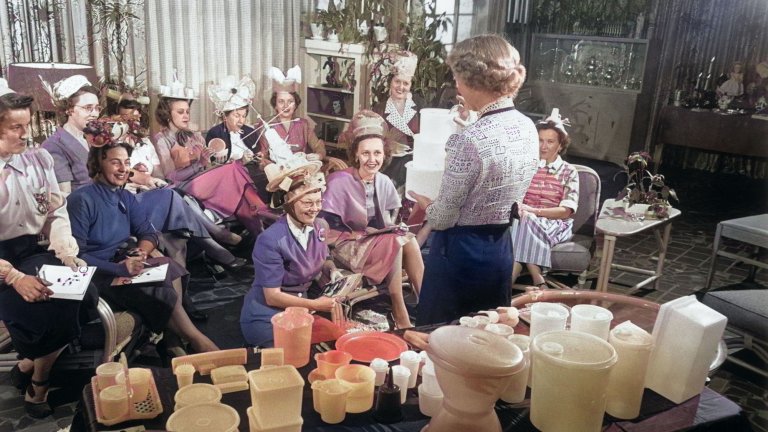 A vintage 1950 photograph woman holds three Tupperware containers while standing in front of a group of women seated in a living room during a Tupperware party