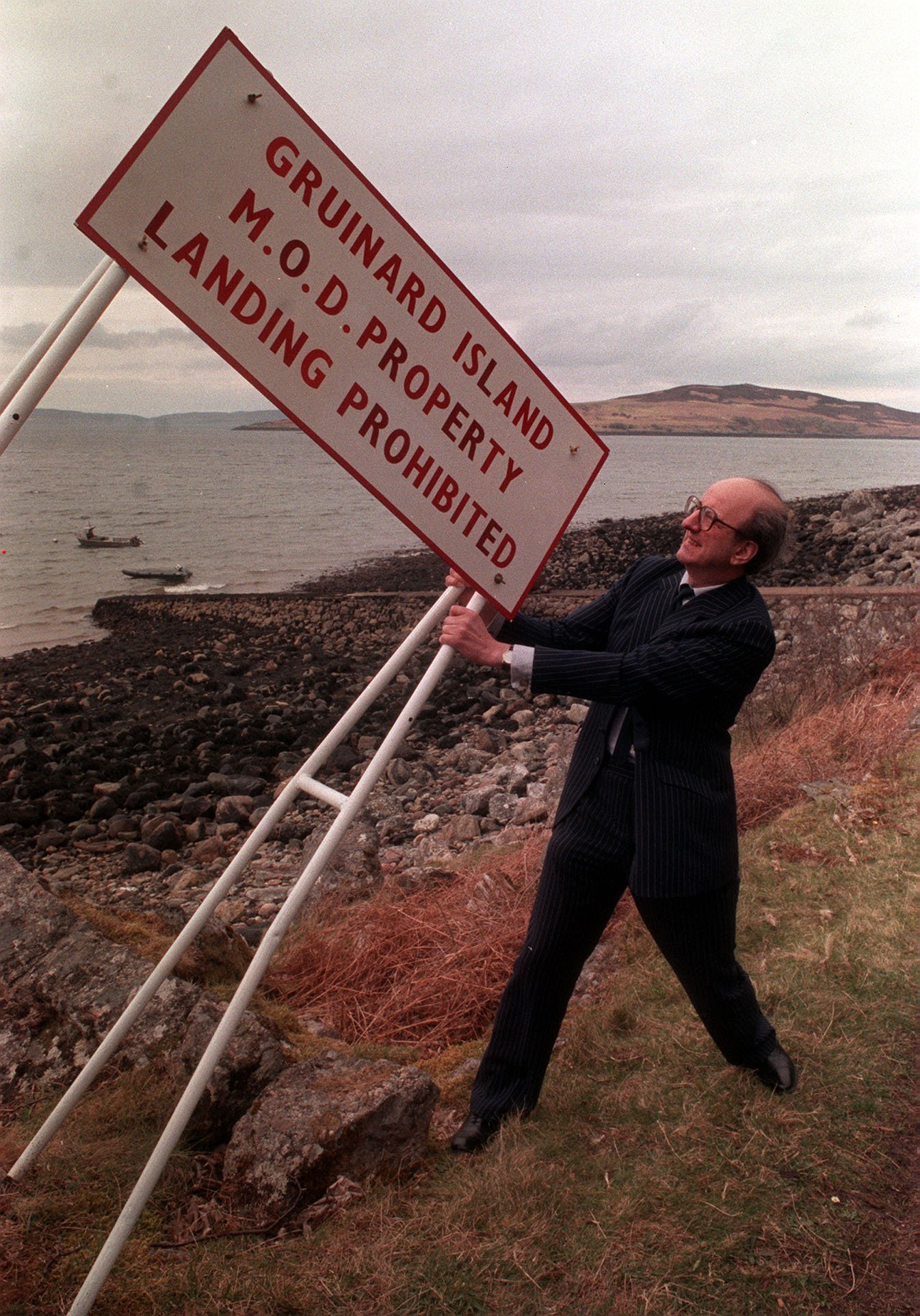A vintage color photograph shows an older man in a suit pulling a sign out of the ground on a remote Scottish coast.