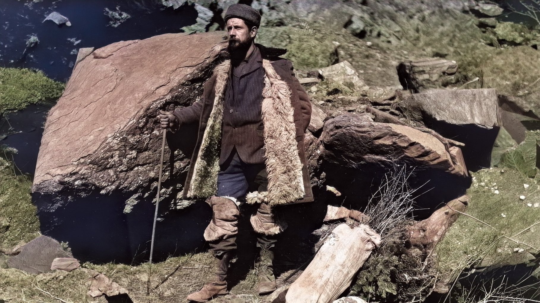 A vintage photograph of a man in a large coat and walking stick in a rocky landscape.