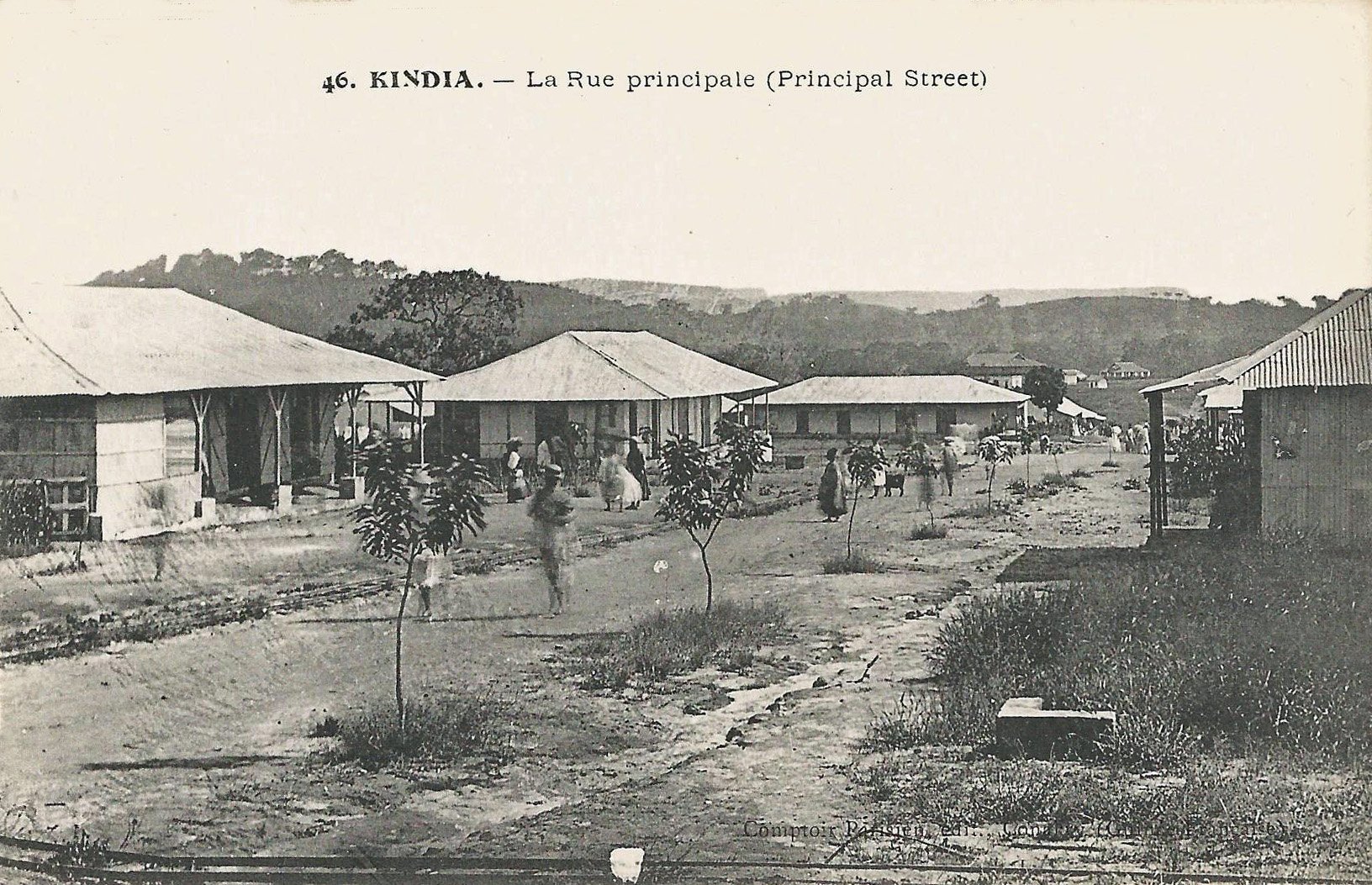 Black and white photograph of a rural street in French Guinea. There are some single floor homes and people walking.