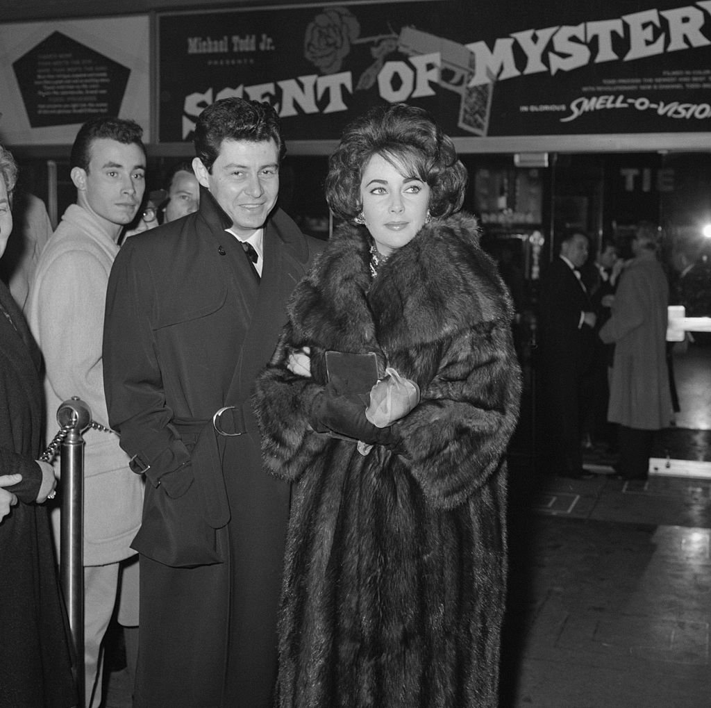 Stylish white woman with black hair in a big fur coat poses for a photograph next to a white man with dark hair.