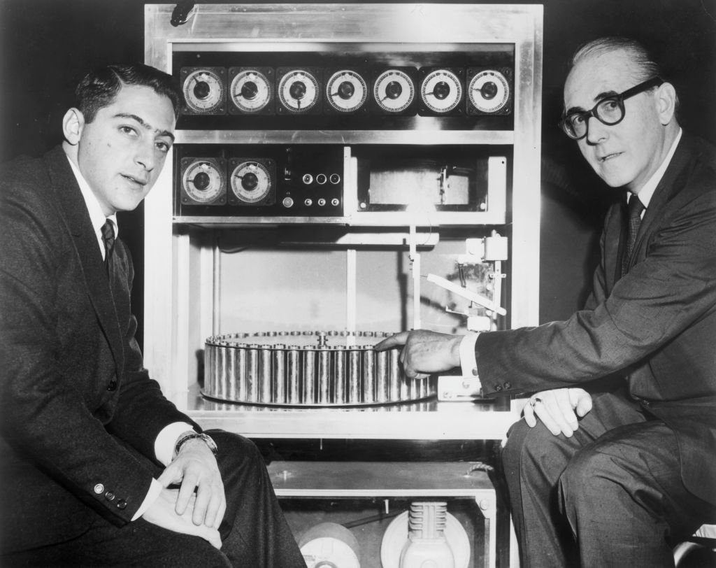 Black and white vintage photograph of two white men sitting next to a machine with a spinning plate of vials and dials. 