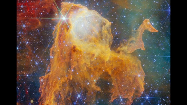 A region of space filled with bright stars and clouds of gas. In the centre, the densest clouds form the shape of a chest with its lid open. The chest appears to glow from within. At its base it breaks apart into long pillars of thick gas. Many of the gas clouds in the background are dark orange globules, while others form large, pale hazes. A few brightly shining stars lie in the foreground, the biggest and brightest in front of the chest’s lid.