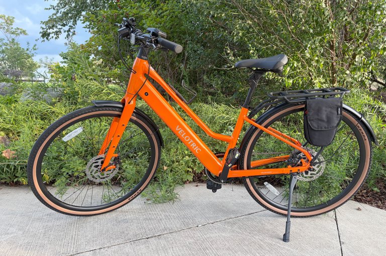 A sunset tangerine Velotric Tempo city commuter e-bike sitting in a green leafy nook
