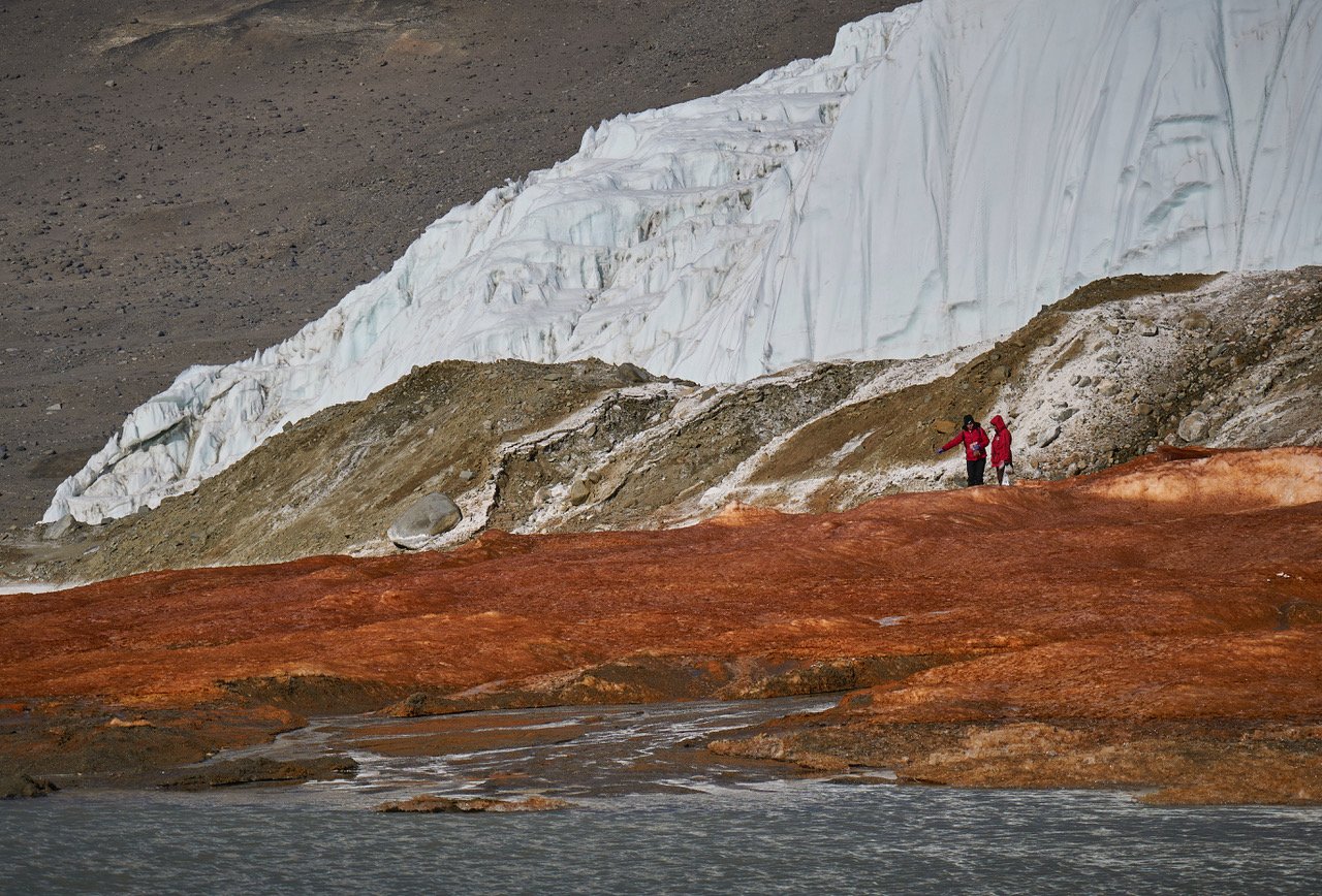 two scientists wearing red jackets standing on redish-orange rock in antarctica